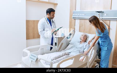 Bedside consultation at modern hospital. Senior woman lying on hospital bed in ward while consultation with friendly Indian doctor and caring nurse Stock Photo