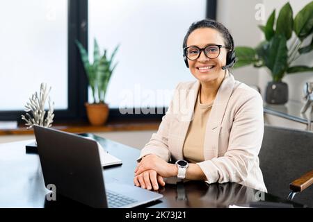 Smiling female call center employee using a headset and laptop for online communication with customers, woman smiling into the camera Stock Photo