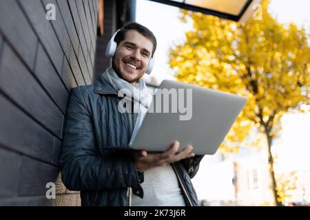 an adult man with a laptop in his hands and headphones leaning against the wall against the backdrop of autumn trees Stock Photo