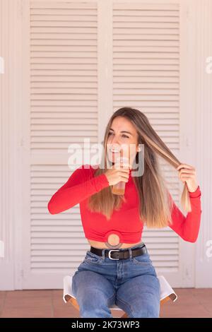 Vertical view of young woman looking to the side while sitting and wearing red and denim outfit spraying moisturising Hair Spray Stock Photo