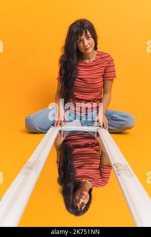 Young, stylish girl with long brown hair sitting on the floor with the mirror laying in front of her. Girls reflection in the mirror, isolated orange background. High quality photo Stock Photo
