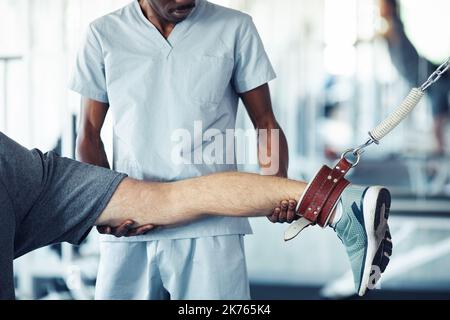 Close-up of therapist in uniform holding leg of patient while he doing stretching exercises in gym Stock Photo