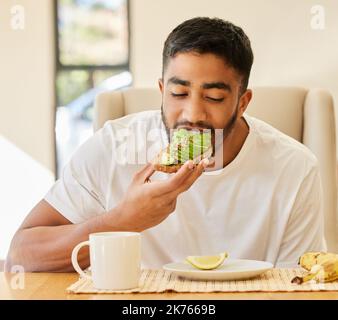 Hmm, avocado on toast is my favourite. a handsome young man sitting alone and having breakfast at home. Stock Photo