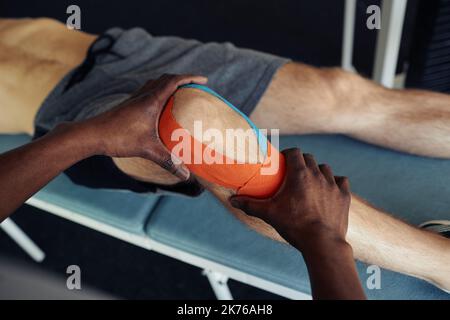 Close-up of therapist applying medical tapes on leg of patient before physical exercises in gym Stock Photo
