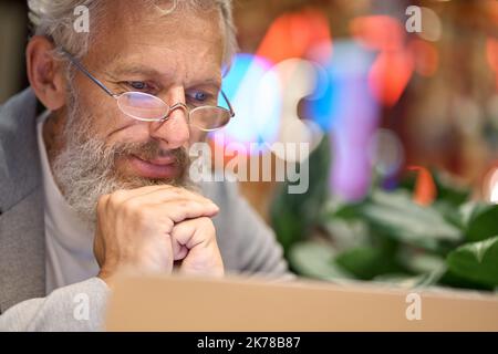 Thoughtful old business man wearing glasses looking at laptop watching webinar. Stock Photo