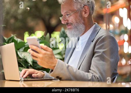Happy older business man holding smartphone using laptop working late. Stock Photo