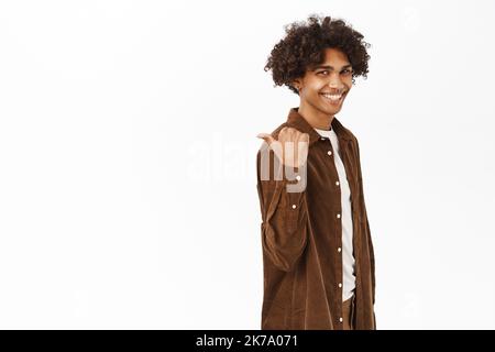 Portrait of handsome young hispanic man pointing finger left, behind his back, showing logo or banner, chart on copy space, standing over white Stock Photo