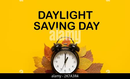 Daylight saving day. Fall Back. Black Alarm clock and autumn leaves and text Daylight saving day on yellow background. Daylight saving time end Stock Photo