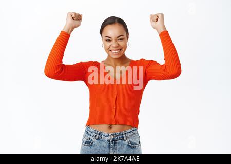 Hooray victory. Excited young woman winning, raising hands up and celebrating, triumphing, smiling with joy, standing over white background Stock Photo