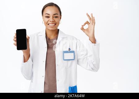 Smiling doctor shows okay sign, mobile phone screen app, medical clinic application, stands over white background Stock Photo