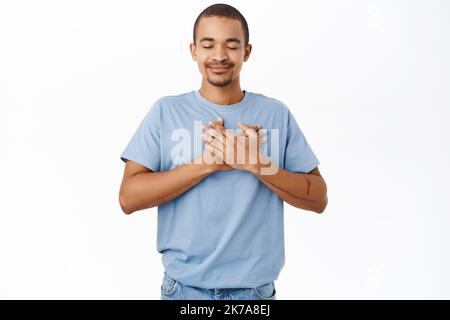 Smiling nice guy holds hands on heart, heartwarming and caring feeling, standing over white background Stock Photo