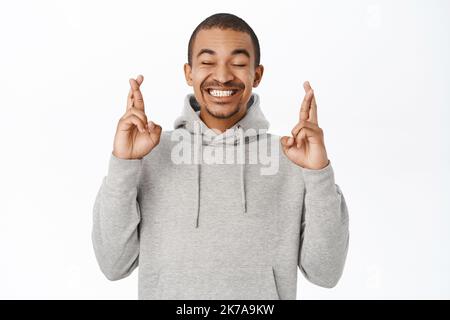 Portrait of hopeful arab guy praying, making wish and holds fingers crossed for good luck, anticipating, standing over white background Stock Photo