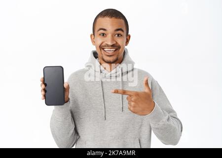 Handsome man shows mobile phone screen, points at telephone and smiles, recommends application, stands over white background Stock Photo