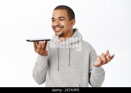 Handsome arab guy record voice message on smartphone, talking into mobile speakerphone, using telephone voice translation, standing over white Stock Photo