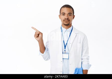 Doctor in white coat pointing finger left. Young intern in clinic showing way, demonstrating promo, standing over white background Stock Photo