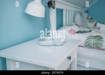 White wireless headphones lie on the bedside table near the bed in hotel room. White wood bedside table. Stock Photo