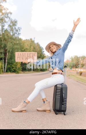 Joyful woman try to stop passing car sitting on suitcase with outstretched cardboard poster on empty highway. Lady in hat escape from city to go Stock Photo