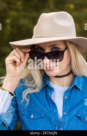 Portrait closeup of beautiful blonde woman in sunglasses and elegant hat on forest background. Stylish traveller in seasonal denim outfit. Autumn Stock Photo