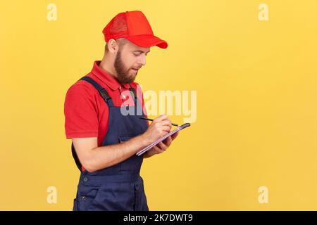 Portrait of serious handyman wearing blue overalls standing with paper notebook in hands, writing down orders, having concentrated expression. Indoor studio shot isolated on yellow background. Stock Photo