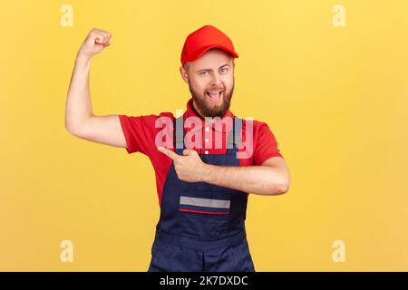 Portrait of proud strong worker man wearing blue uniform and red cap standing with raised arms and pointing at his biceps, showing his power. Indoor studio shot isolated on yellow background. Stock Photo