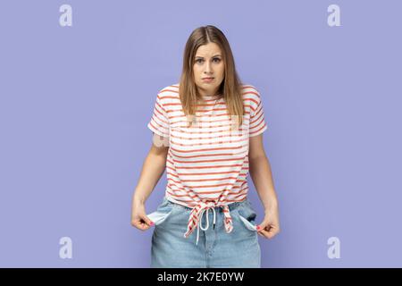 Unemployment and bankruptcy. Portrait of blond woman wearing striped T-shirt turning out empty pockets, worried about debts, no cash for living. Indoor studio shot isolated on purple background. Stock Photo