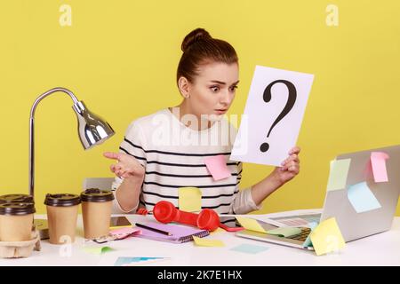 Surprised astonished woman looking at laptop monitor, holding paper with question mark, thinks about tasks, covered with sticky notes. Indoor studio studio shot isolated on yellow background Stock Photo