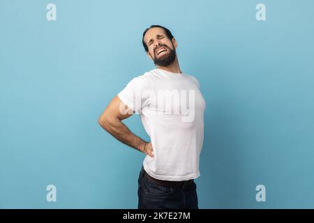 Portrait of man with beard wearing white T-shirt holding hands on back with painful grimace, suffering from backache or kidney inflammation. Indoor studio shot isolated on blue background. Stock Photo