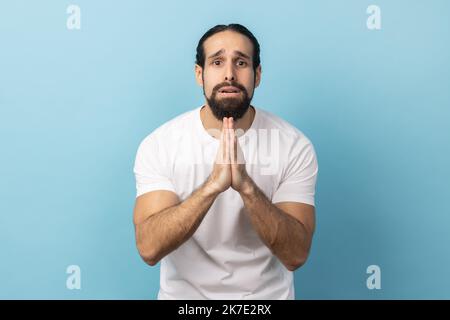 Please, I'm begging forgive. Portrait of upset worried man with beard wearing white T-shirt looking with imploring desperate grimace, praying for help. Indoor studio shot isolated on blue background. Stock Photo