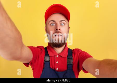 Surprised amazed worker man with wearing red cap and blue uniform taking selfie, looking at camera with big eyes POV, point of view of photo. Indoor studio shot isolated on yellow background. Stock Photo