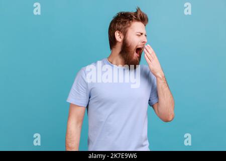 Need rest. Portrait of bearded man yawning and dreaming of sleep, feeling drowsy, inefficient to do job, laziness and lack of energy. Indoor studio shot isolated on blue background. Stock Photo