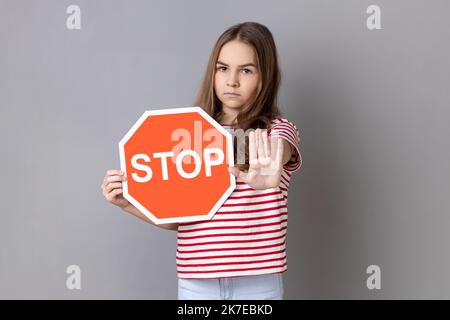 Little girl in T-shirt covering face with octagonal Stop symbol, showing red traffic sign and ban gesture with palm, warning of forbidden way. Indoor studio shot isolated on gray background. Stock Photo