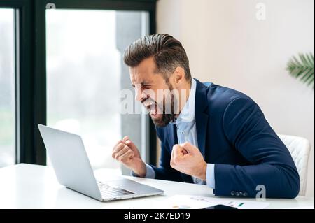 Angry annoyed frustrated caucasian business man, sitting at workplace with laptop, screaming, gesturing with fists, failed deal, disappointed with project result, experiencing negative emotions Stock Photo