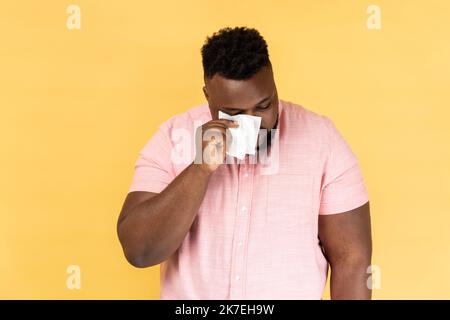 Portrait of man wearing pink shirt hiding face in hand with white handkerchief, crying, feeling stressed worried facial expression. Indoor studio shot isolated on yellow background. Stock Photo