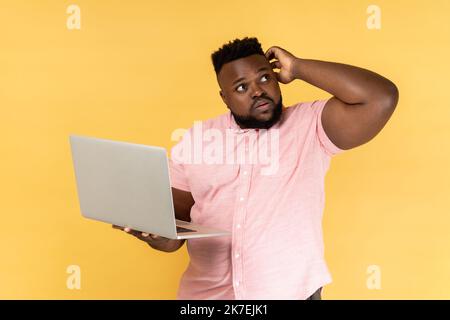 Portrait of thoughtful bearded man wearing pink shirt holding laptop and thinking over startup strategy with serious doubtful expression. Indoor studio shot isolated on yellow background. Stock Photo