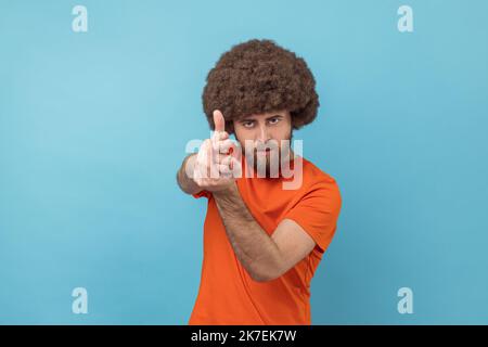 Portrait of serious man with Afro hairstyle wearing orange T-shirt holding fingers pretending holding gun, aiming enemy, self-defense. Indoor studio shot isolated on blue background. Stock Photo