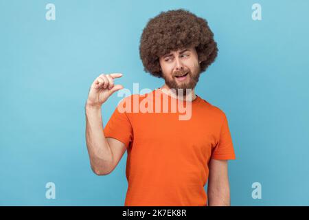 Not very much. Portrait of man with Afro hairstyle wearing orange T-shirt shapes little gesture, demonstrates something very tiny, measures small size. Indoor studio shot isolated on blue background. Stock Photo
