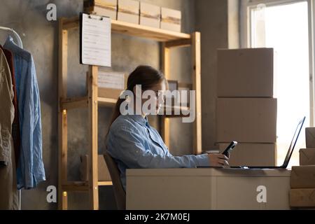 Female warehouse worker manager, small business owner holding phone have retail package parcel boxes checking commercial shipping delivery order on Stock Photo