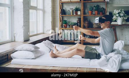 Happy young man in virtual reality glasses is sitting on bed and moving hands as if he is driving car turning steering wheel, guy is enjoying new experience and laughing. Stock Photo