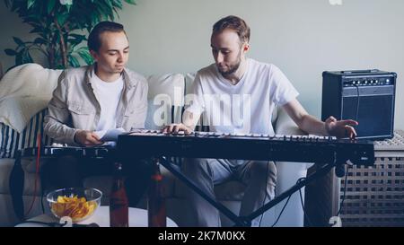 Young bearded keyboarder is adjusting musical instrument then playing looking at music sheet and talking to his friend guitarist who is sitting on sofa in studio during rehearsal. Stock Photo