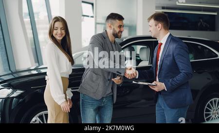 Young bearded man is buying car for his attractive wife shaking hands with manager and getting key Stock Photo