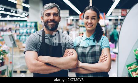 Portrait of two supermarket employees attractive young people in aprons standing inside shop, smiling and looking at camera. Shelves with food and drinks are visible. Stock Photo