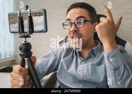 Angry sad upset man in blue wear emotionally talking on mobile phone over white office background. indoor. Live broadcast via the Internet. Concept Stock Photo