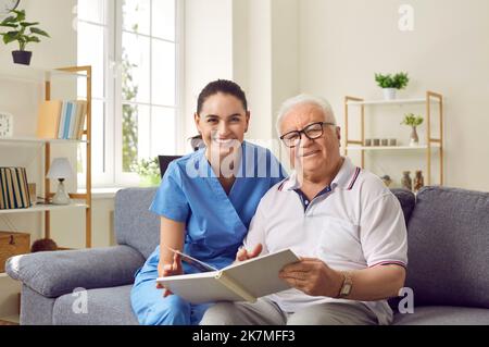 Happy senior man and young female nurse sitting on couch and reading book together Stock Photo