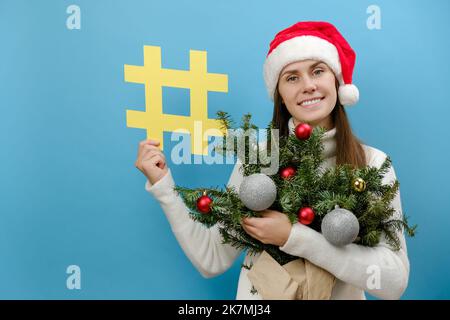Portrait of pretty young caucasian female in Christmas red hat holding bouquet of spruce branches and hashtag symbol, posing isolated on blue backgrou Stock Photo