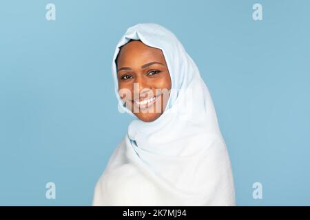 Portrait of happy african american muslim woman in hijab looking and smiling at camera, standing over blue background Stock Photo