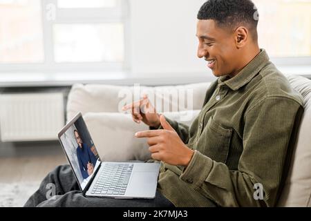 Side view of the concentrated male student involved virtual meeting, webinar. Black man making video call on the laptop, sitting on the sofa at home, talking and gesturing, while explaining something Stock Photo