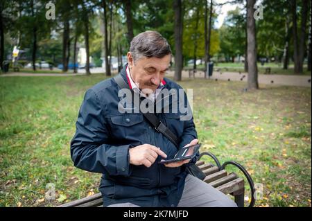 Portrait of a smiling elderly man sitting on a bench in a public park. The old man is resting in the fresh air and talking on the phone. A man is look Stock Photo