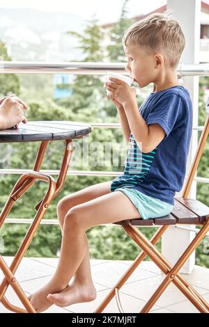 Preschooler boy drinks warm milk from mug sitting on chair in early morning. Excited child enjoys summer holidays drinking beverage on hotel balcony Stock Photo