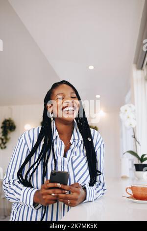 Portrait of a young businesswoman laughing happily while holding a smartphone in a coffee shop. Cheerful young black businesswoman enjoying her coffee Stock Photo