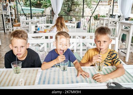 Brothers sit at wooden table in restaurant drinking lemonade from glasses using straws. Siblings enjoy going together to restaurant to taste meals Stock Photo